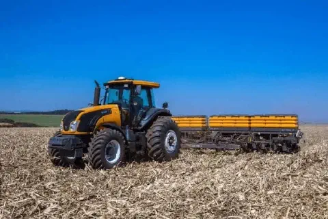 trator de grande porte, de cor amarela e preta, operando em uma lavoura sob um céu azul claro e sem nuvens.