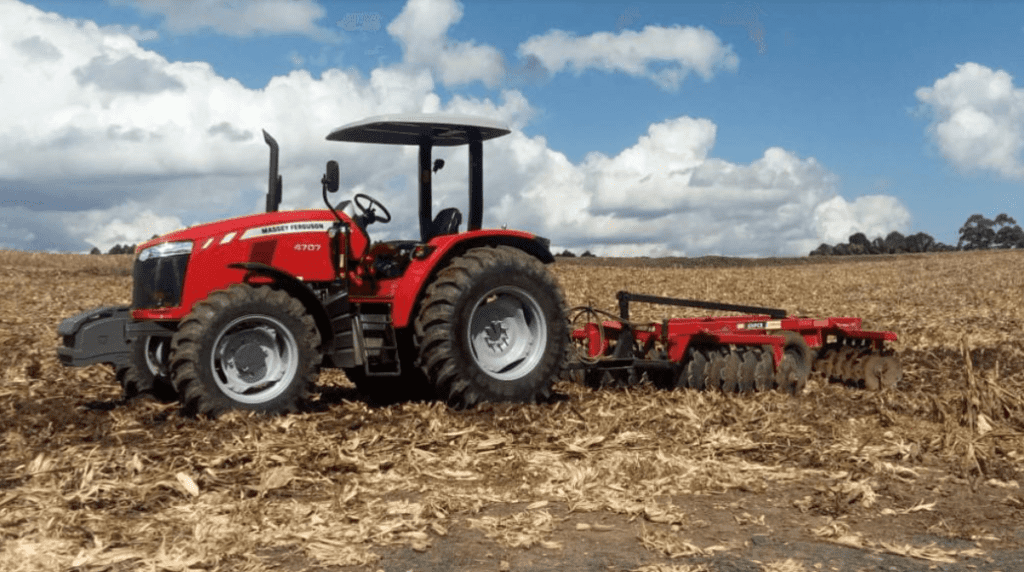 trator vermelho da marca Massey Ferguson, modelo 4707, parado em um campo agrícola após a colheita. O trato