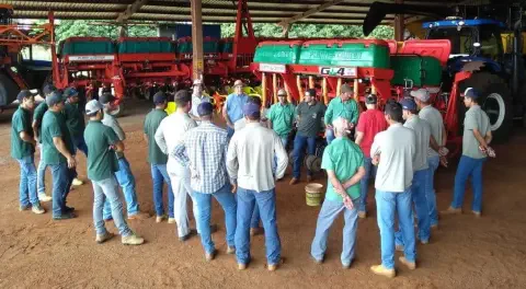 grupo de agricultores e operadores de máquinas reunidos em um grande galpão de fazenda, com piso de terra
