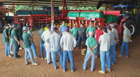 grupo de agricultores e operadores de máquinas reunidos em um grande galpão de fazenda, com piso de terra