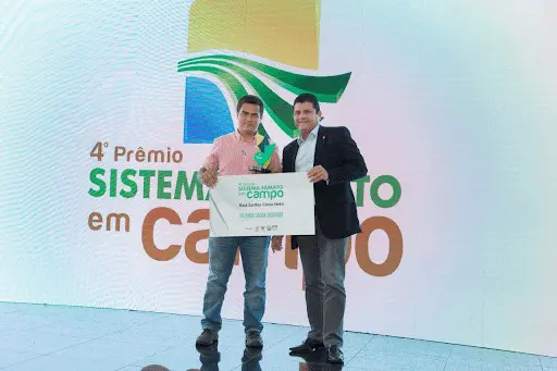 imagem retrata um momento de premiação em um evento do agronegócio. Dois homens estão em um palco, em frente a um grande pa