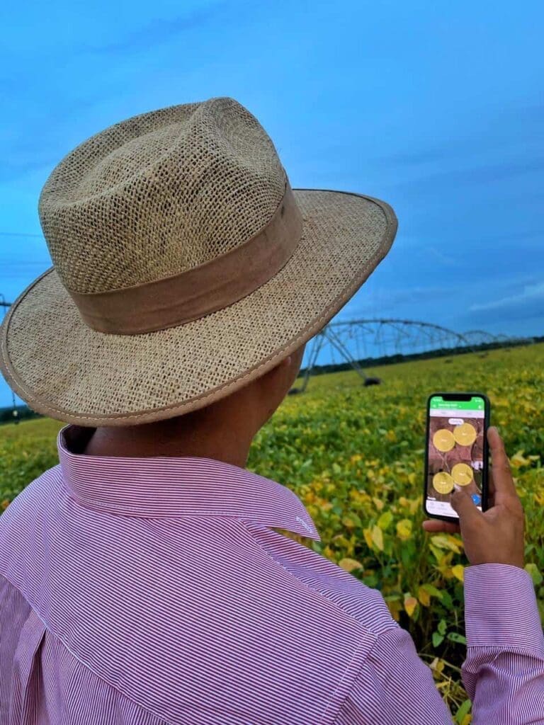 Produtor rural com camisa rosa e chapéu bege, apontando celular para plantação, registrando dados no App Aegro.