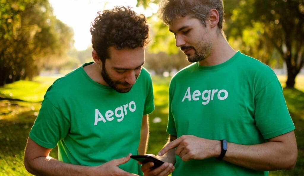 Dois fundadores do Aegro usando camiseta verde com logo da Aegro, olhando para o celular, com árvores e gramado ao fundo.